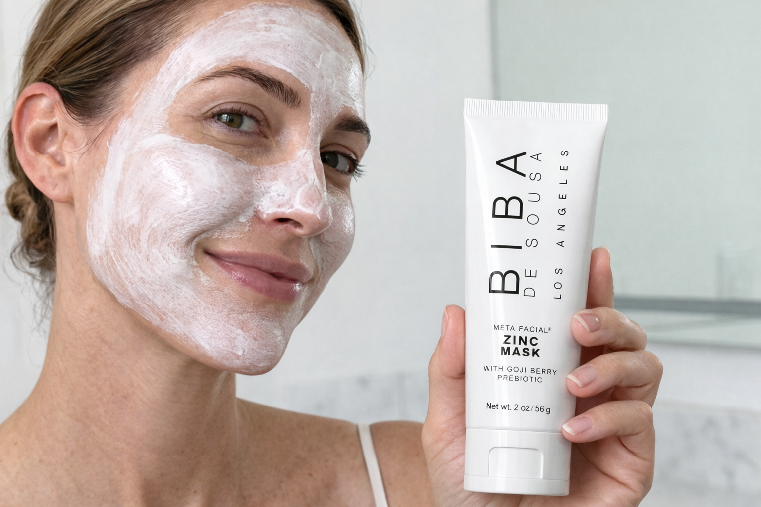 Woman wearing a white zinc facial mask holding Biba de Sousa Los Angeles Zinc Mask with Goji Berry Prebiotic in a softly lit bathroom setting.
