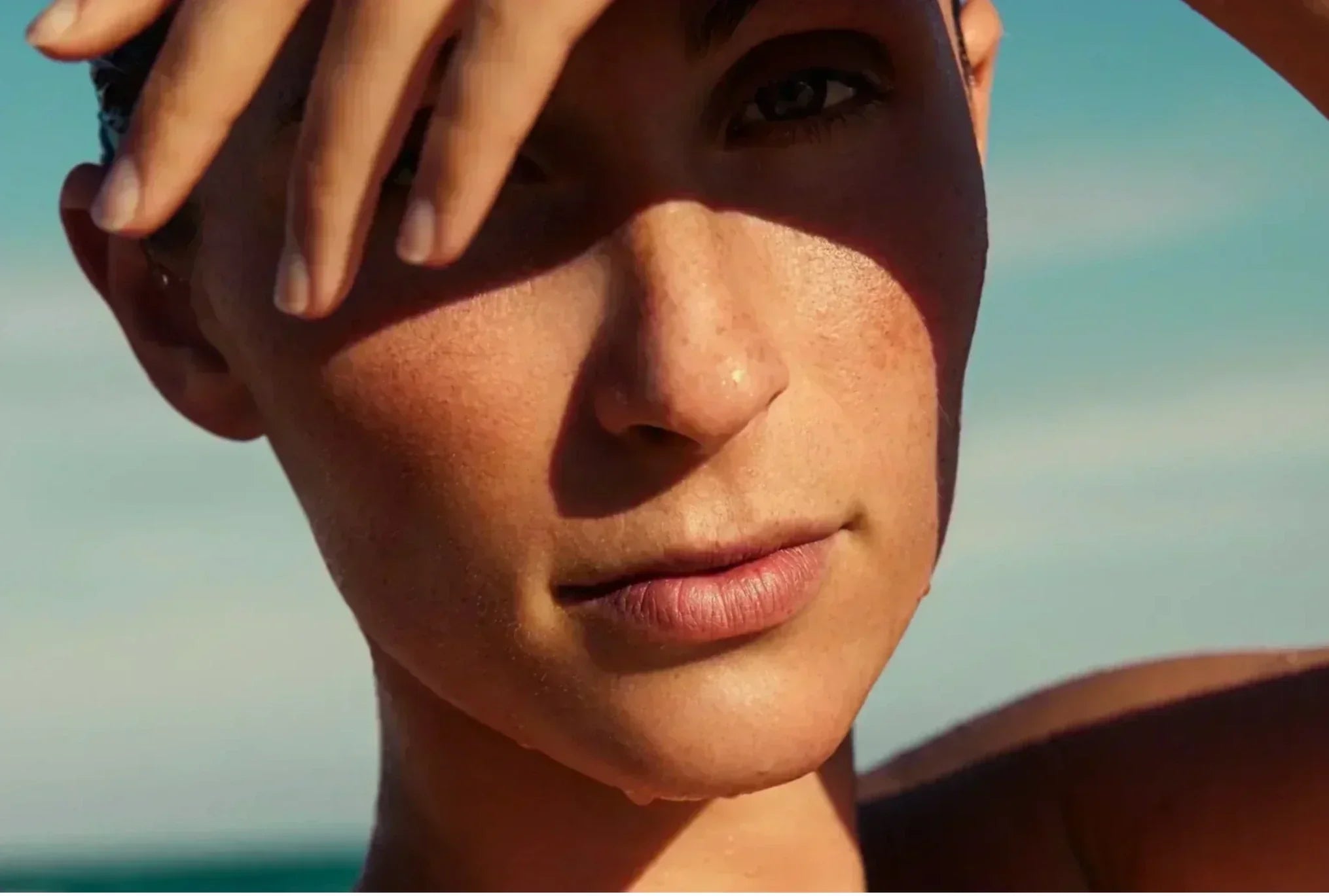 Woman shielding her face from the sun outdoors, illustrating healthy summer skin and the importance of daily sun protection.