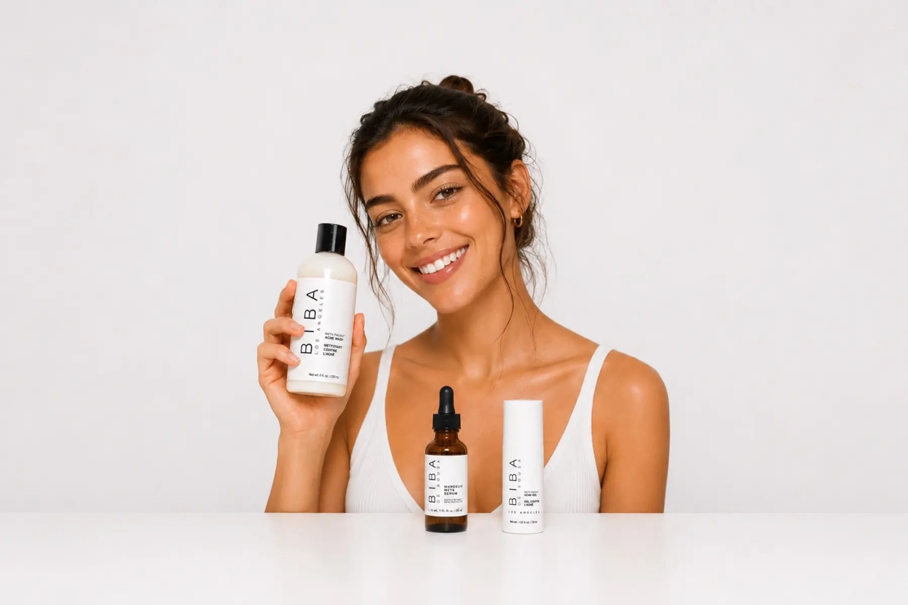 Smiling woman holding Biba Acne Wash with Mandelic Blemish Serum and Acne Gel displayed in front on a light gray background, representing a complete acne skincare routine for clearer, healthier-looking skin.