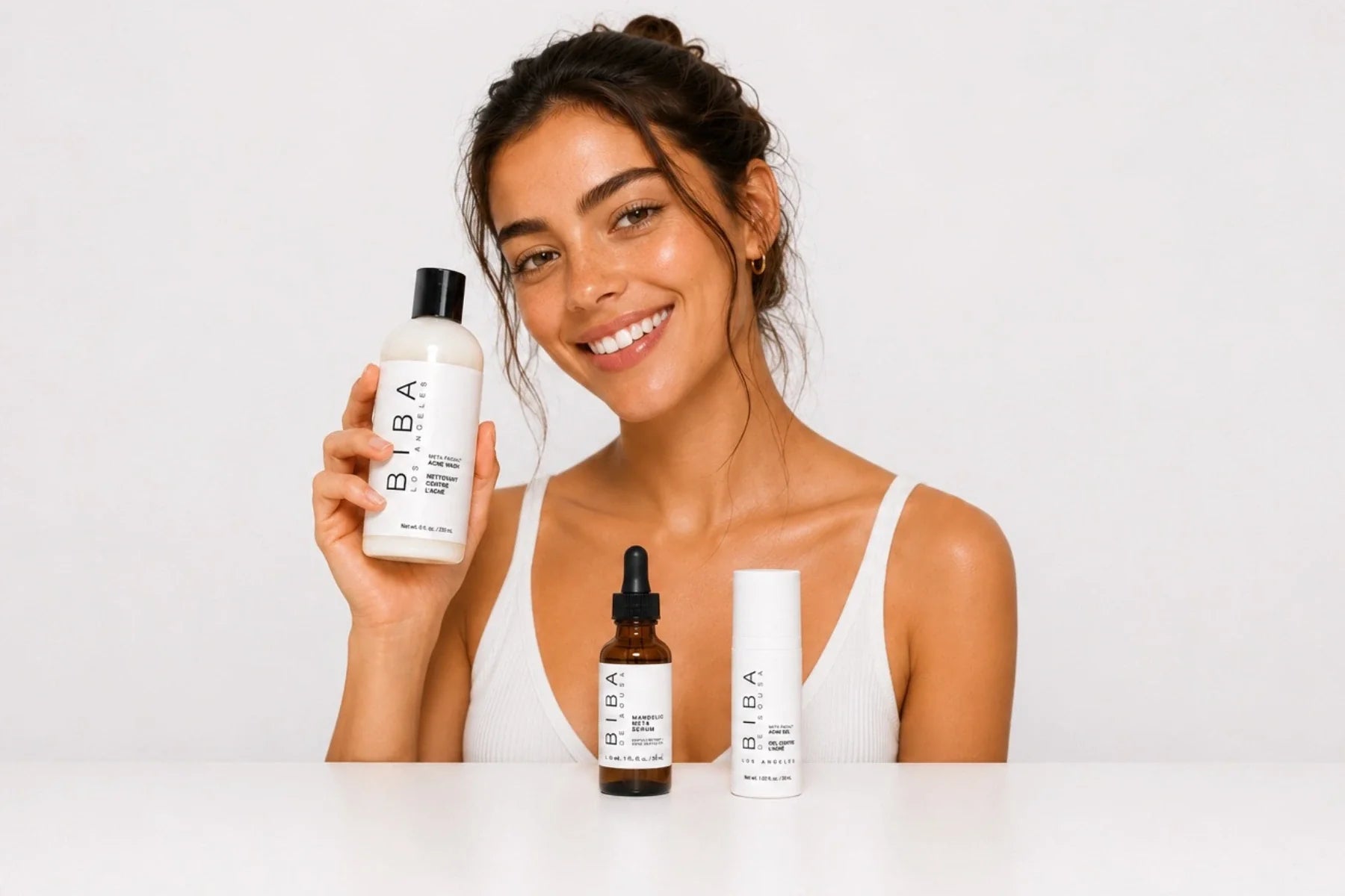 Woman holding BIBA Acne Wash with Mandelic Meta Serum and Acne Gel on a light gray background, representing the BIBA Los Angeles acne skincare collection.