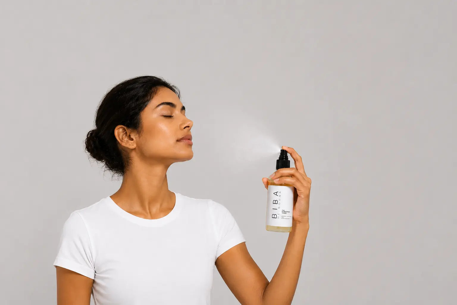 Woman with eyes closed misting Biba Hydrating Toner onto her face against a light gray background with generous negative space, highlighting hydration, antioxidant support, and refreshed, healthy-looking skin.