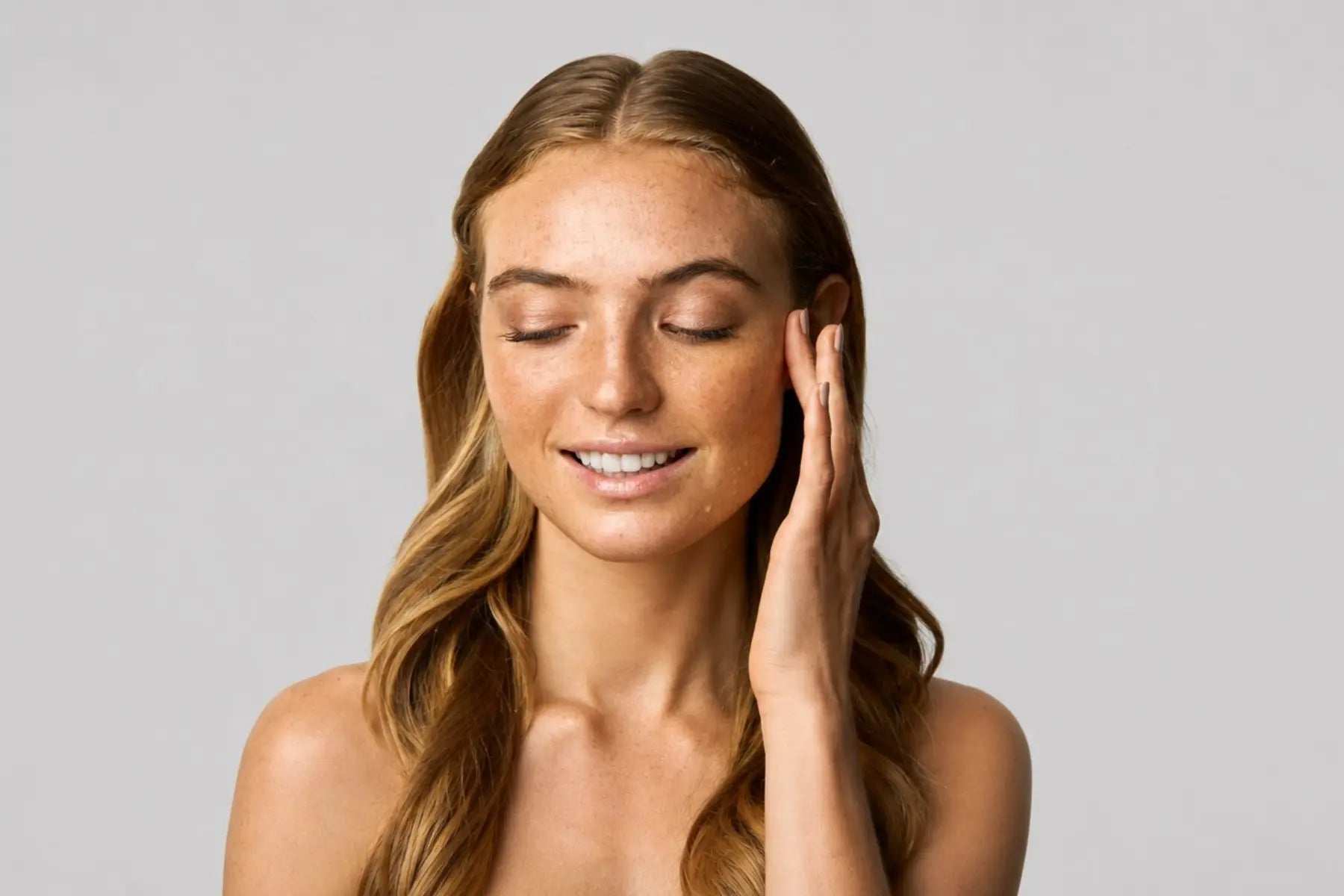 Young woman with visible hyperpigmentation and freckles gently touching her face with eyes closed against a light gray background, representing skincare for uneven skin tone and hyperpigmented skin.