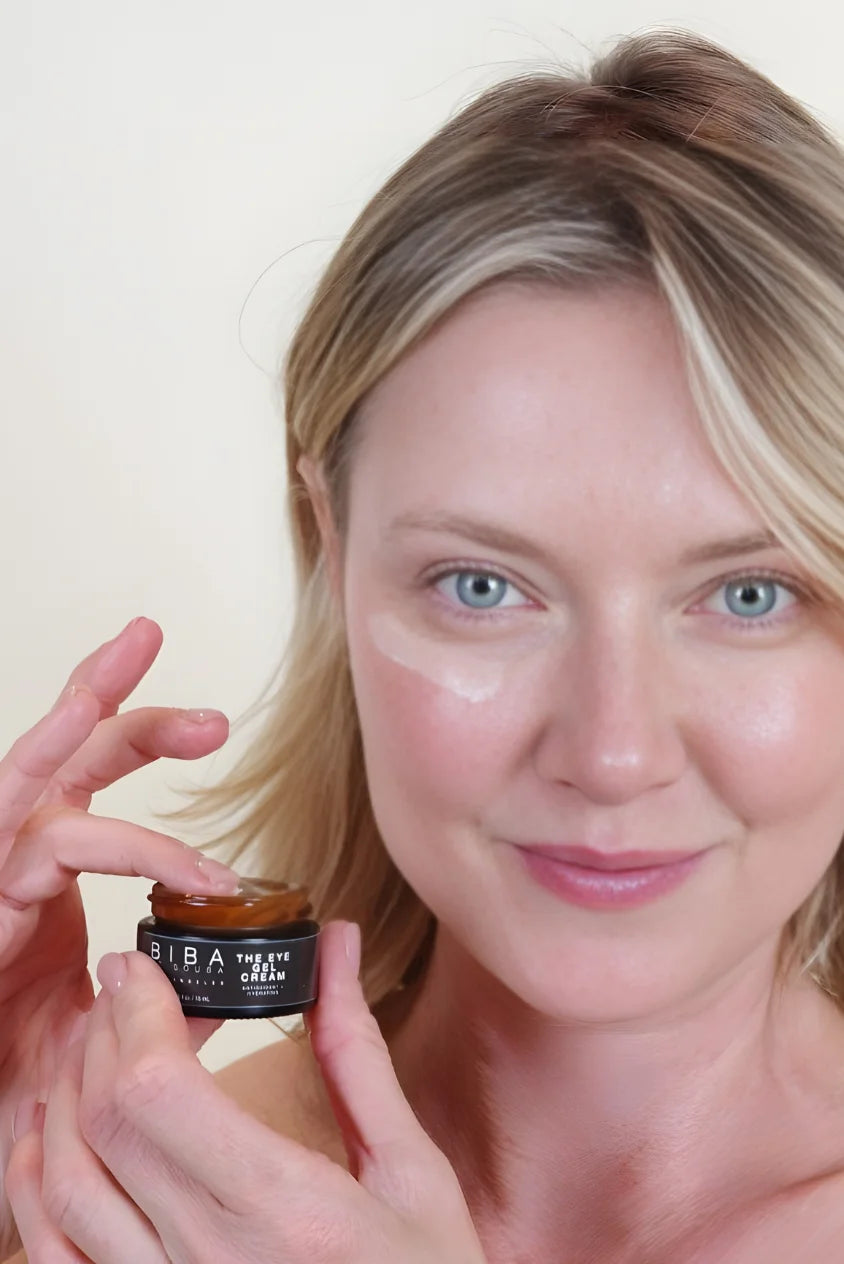 Woman applying Biba Los Angeles Eye Gel Cream under her eye, holding a small open jar of the product, showing a lightweight gel texture on a natural, makeup-free face.