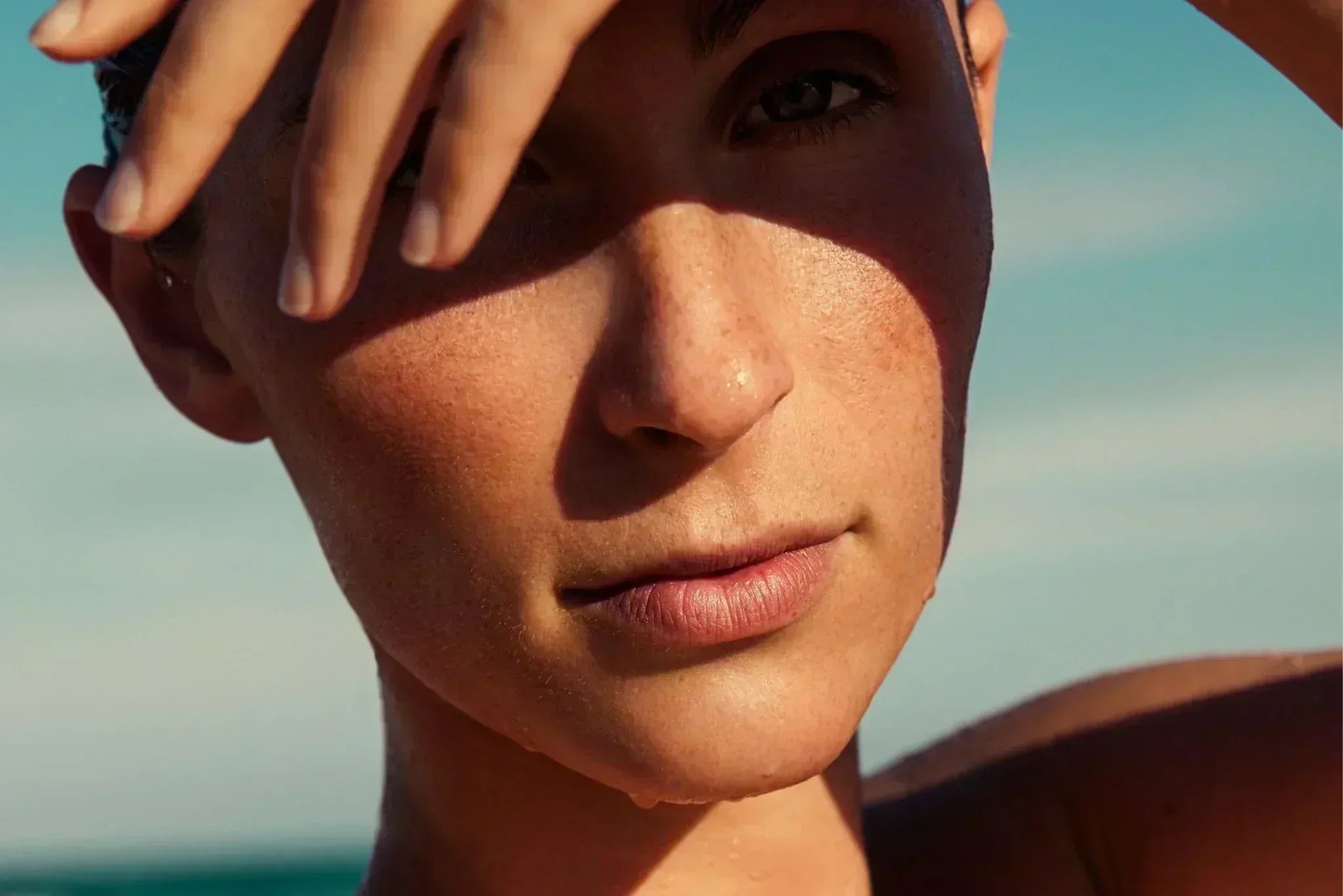  Woman shielding her face from the sun, showcasing natural glowing skin outdoors—illustrating summer skincare and sun protection.