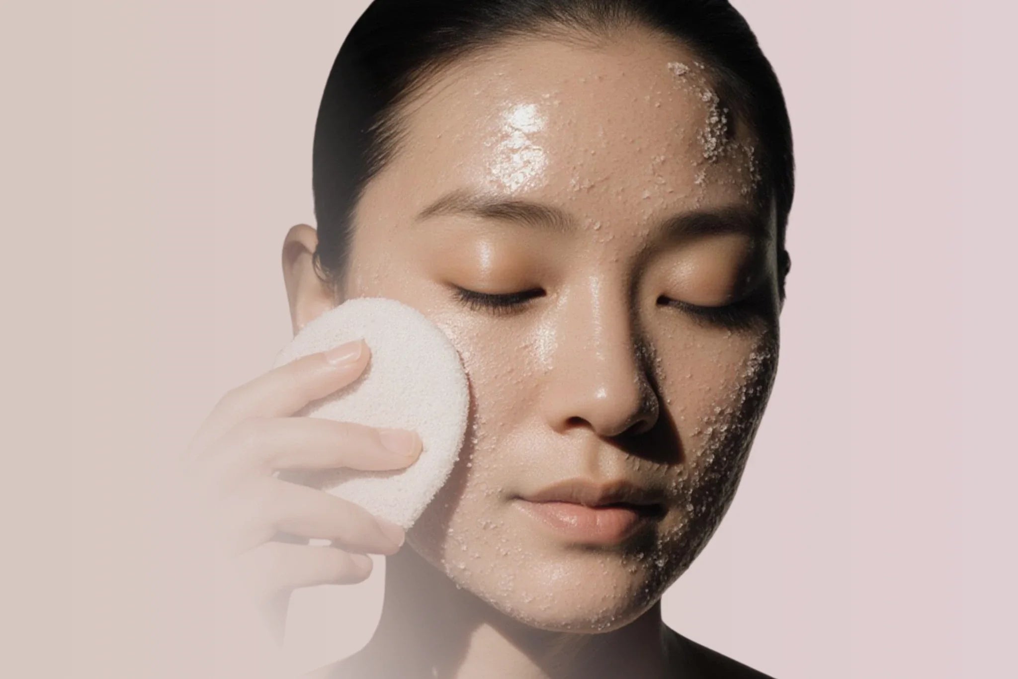 Model with eyes closed gently pressing a soft cleansing pad to her cheek, showing exfoliating particles on dewy skin against a soft neutral background.