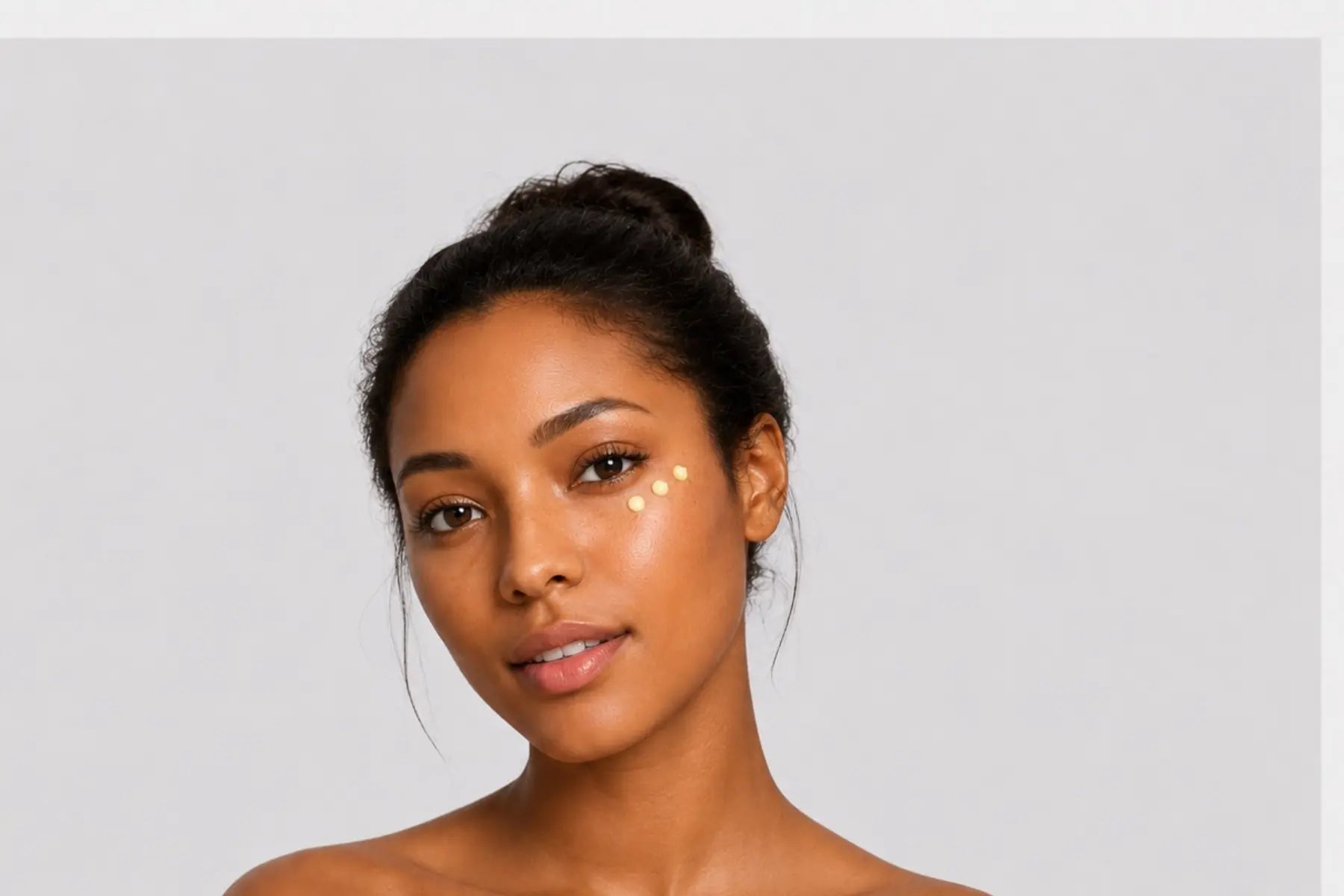 Young woman with glowing skin and three eye cream dots beneath her eye against a light gray background, illustrating the BIBA Eye Cream Collection.