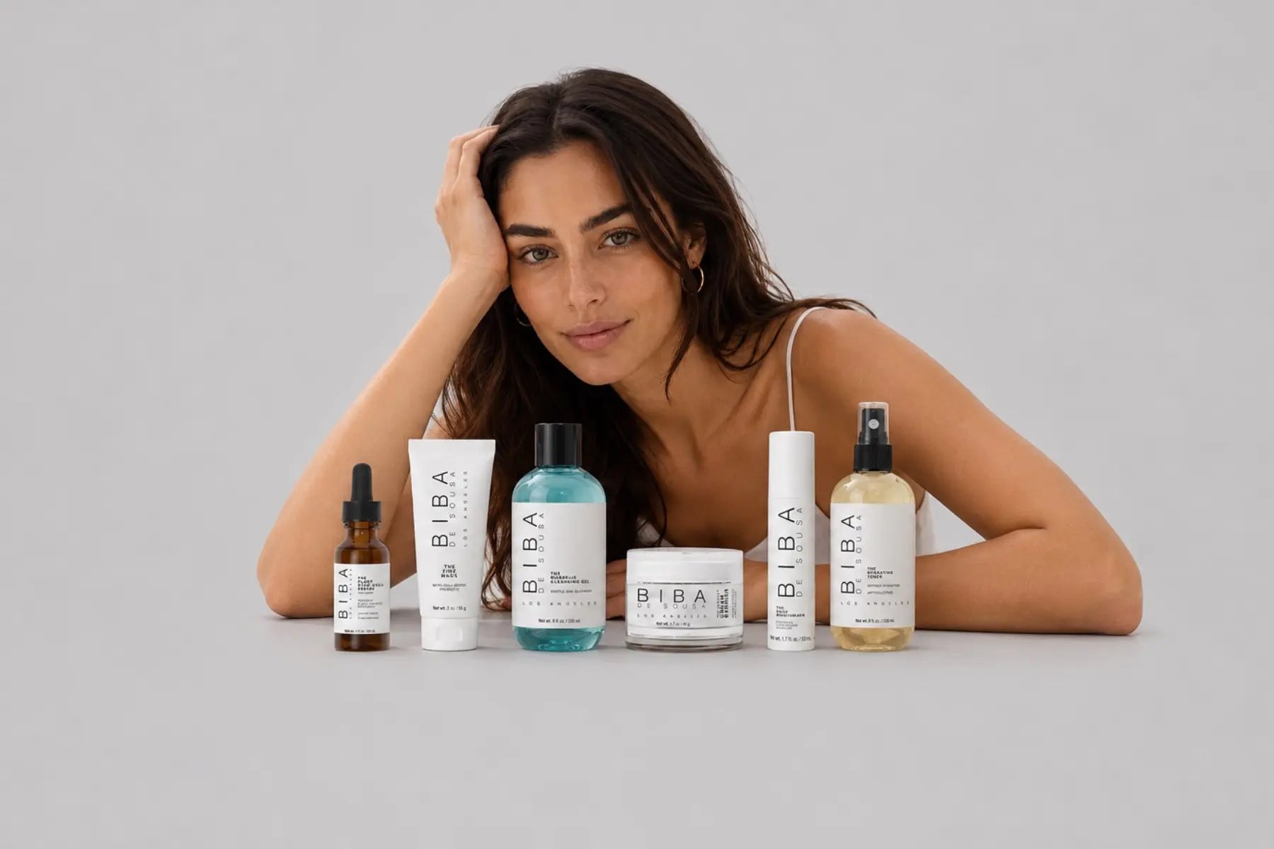 Woman with healthy glowing skin behind the BIBA Los Angeles skincare collection featuring Plant Stem Cell Serum, Zinc Mask, Mandelic Cleansing Gel, Cream Barrier, Daily Moisturizer, and Hydrating Toner on a light gray background.