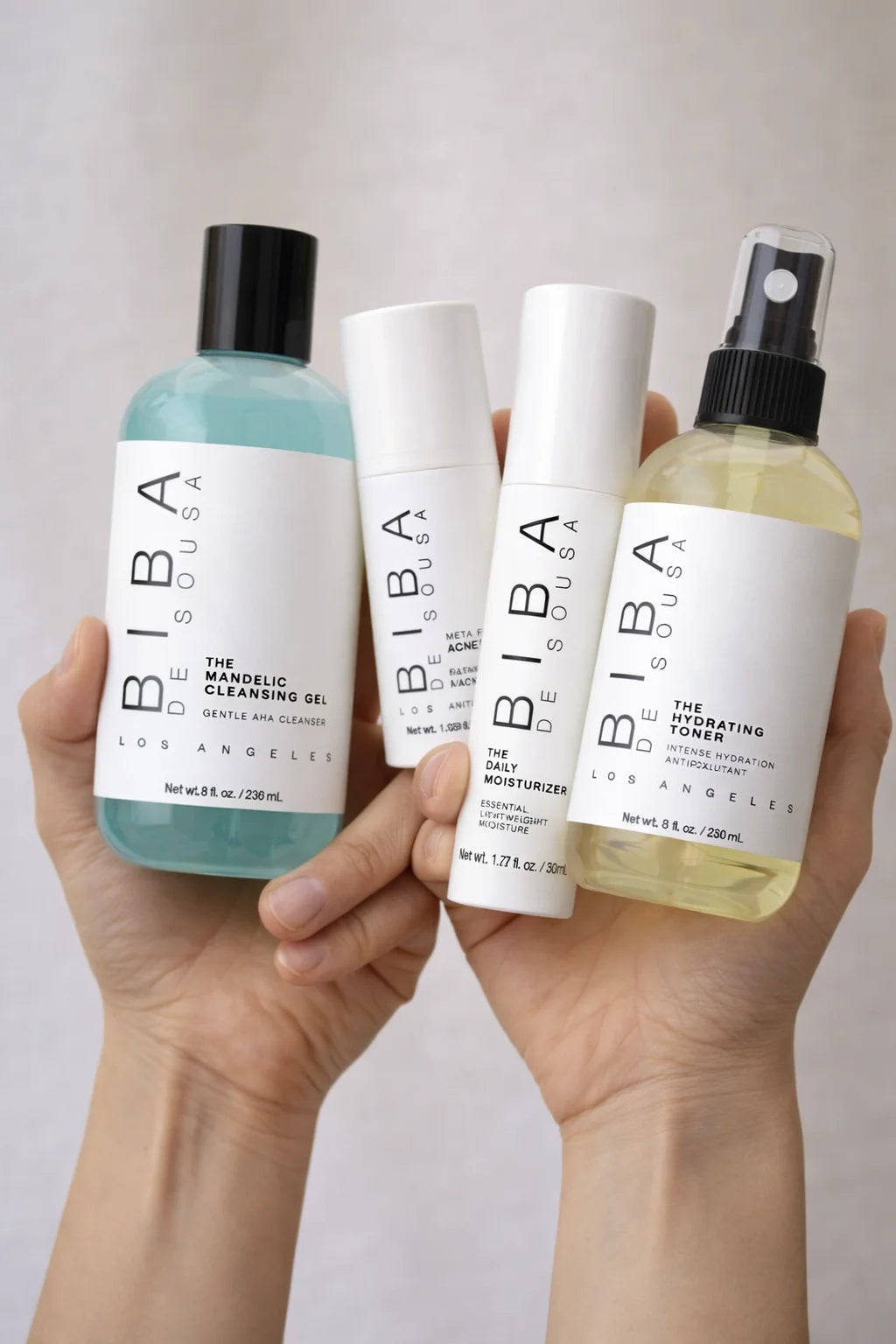 Two hands holding four Biba de Sousa Los Angeles skincare products against a soft neutral background, including a blue Mandelic Cleansing Gel bottle, a white Acne Gel, a white Daily Moisturizer, and a yellow-toned Hydrating Toner spray, all with clean, minimal packaging and labels clearly visible.
