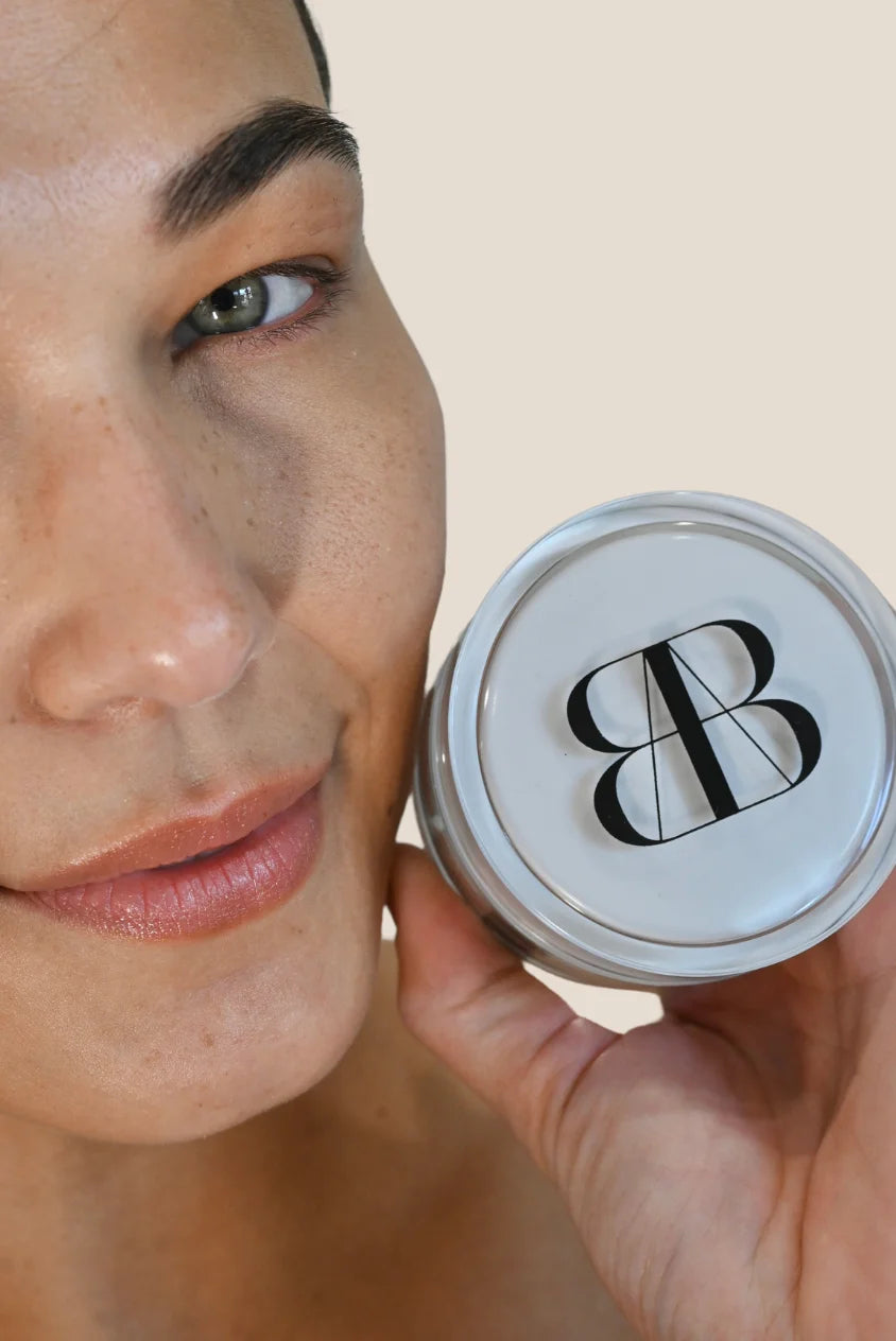 Close-up of a model holding BIBA Los Angeles Cream Barrier moisturizer jar beside her face, showing the product lid and natural skin texture.