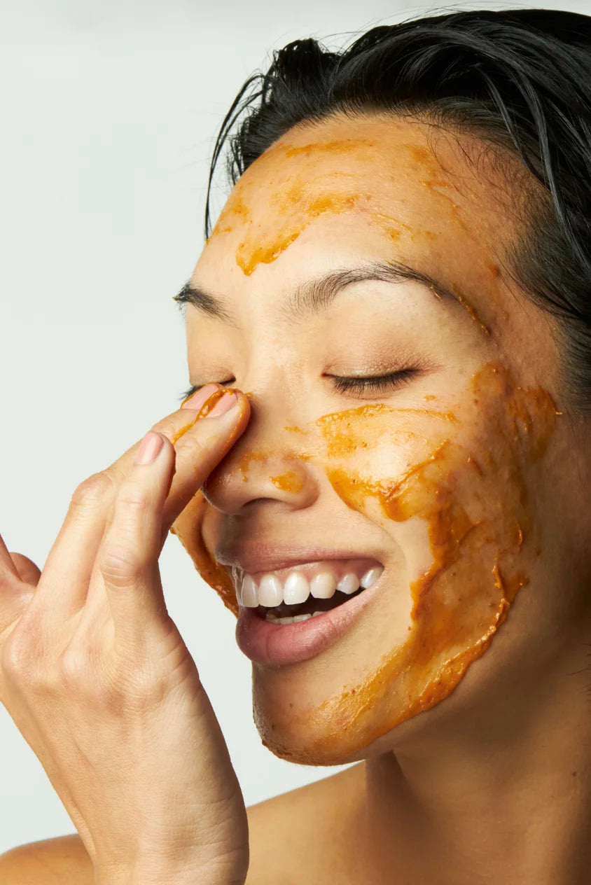 Woman applying a pumpkin enzyme exfoliating mask to her face, smiling with eyes closed against a clean, neutral background.