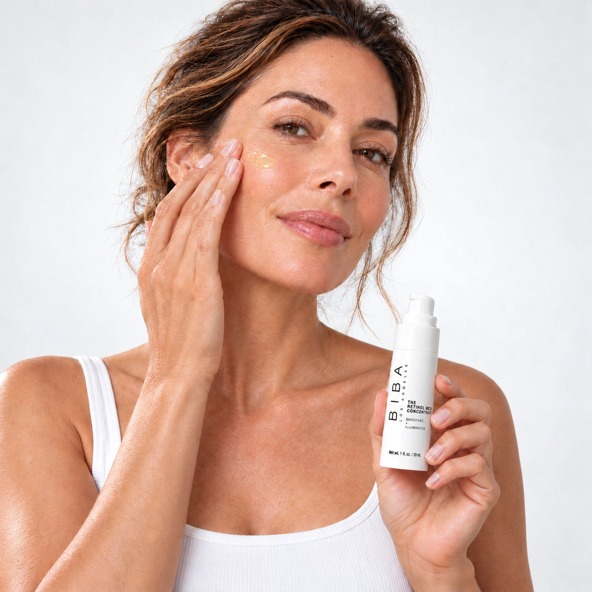 Woman applying pale yellow serum to her cheek while holding a BIBA Los Angeles Retinol Meta Concentrate bottle against a white background.
