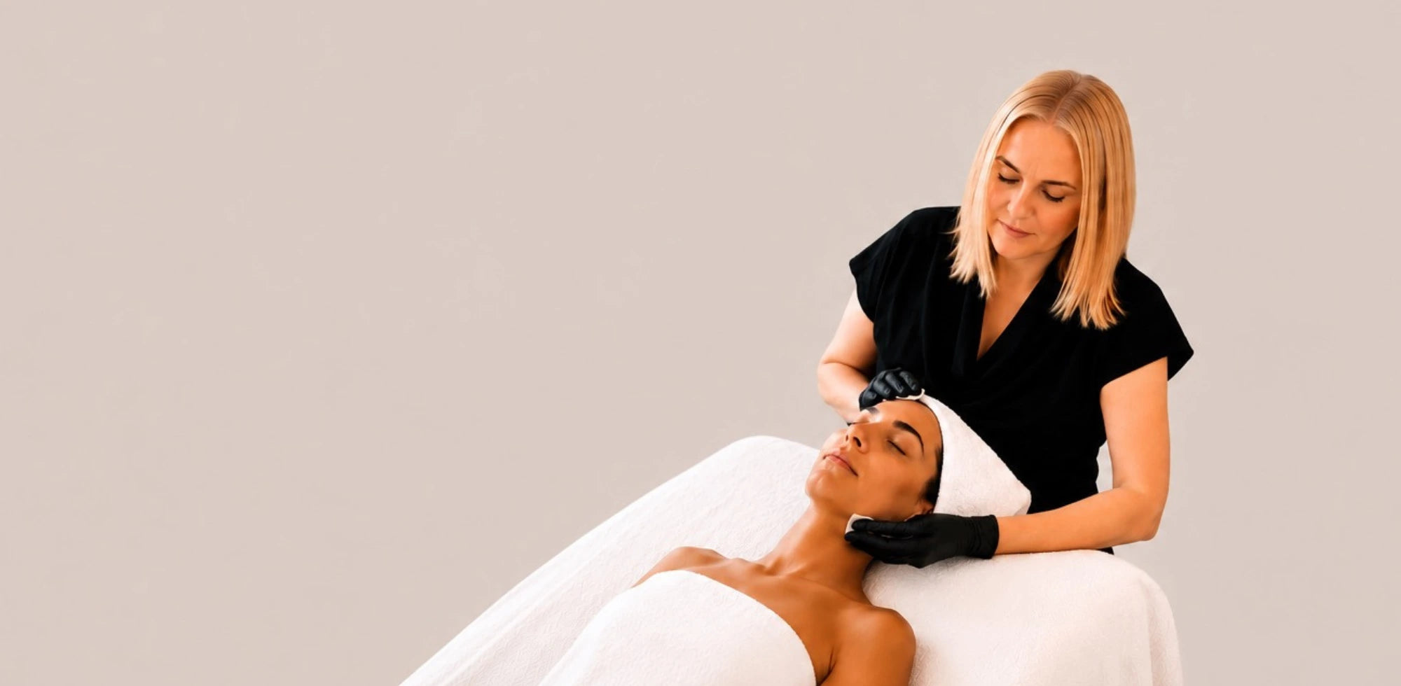 A woman in a black scrub top and gloves performs a facial treatment on a client lying on a white spa table against a solid light gray background.